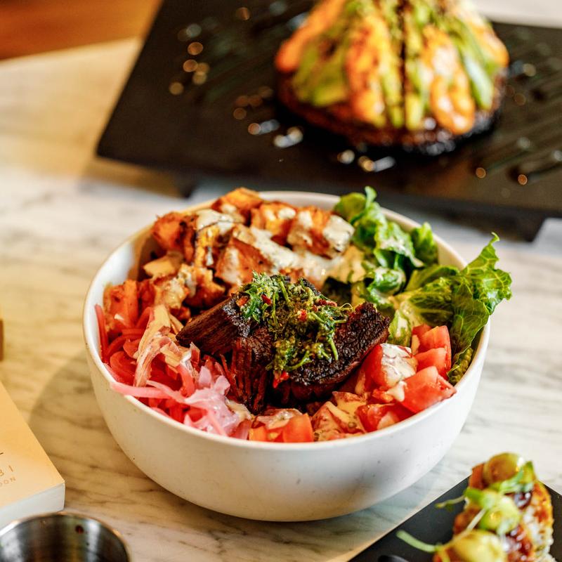 A white bowl of churrasco steak over sweet potato, romaine, tomato and pickled onions with chimichurri and cashew crema.